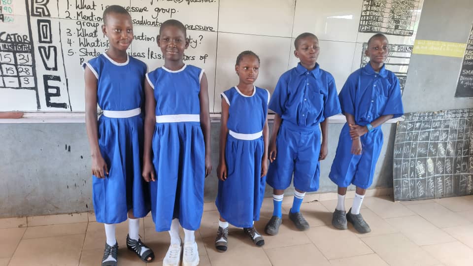 Five children wearing blue uniforms in front of a classroom chalkboard
